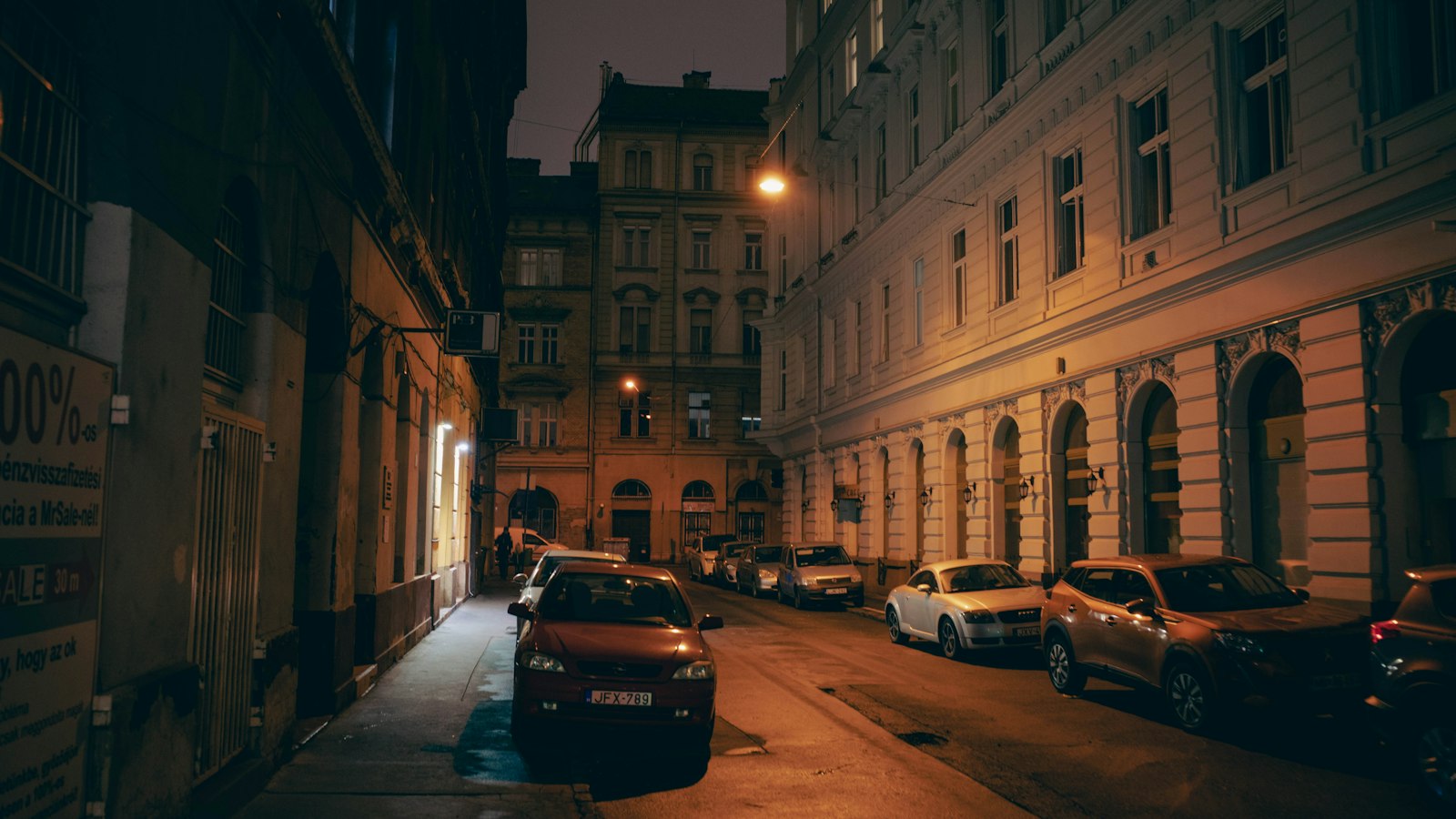 Ulica w centrum miasta nocą, wzdłuż chodnika zaparkowane samochody